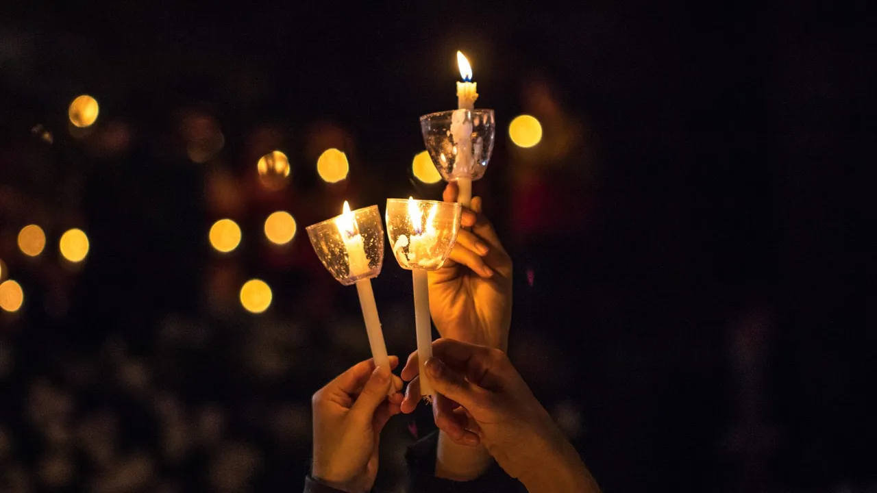 Velas acesas seguradas por mãos em um ambiente escuro com luzes ao fundo